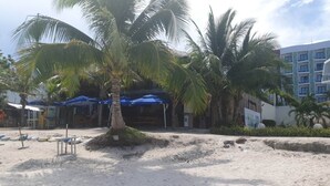 A beachside area with palm trees, a sandy beach, and a building with multiple balconies. - Alona Beach Poolside Hotel (Panglao)