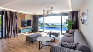 Een moderne woonkamer met een grote bank, een salontafel en een aan de muur bevestigde televisie. Door grote ramen is een zwembad te zien.