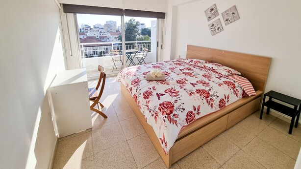 A bedroom with a bed, a desk, a chair, and a balcony with a view of buildings. - City View Rooms (Larnaca)