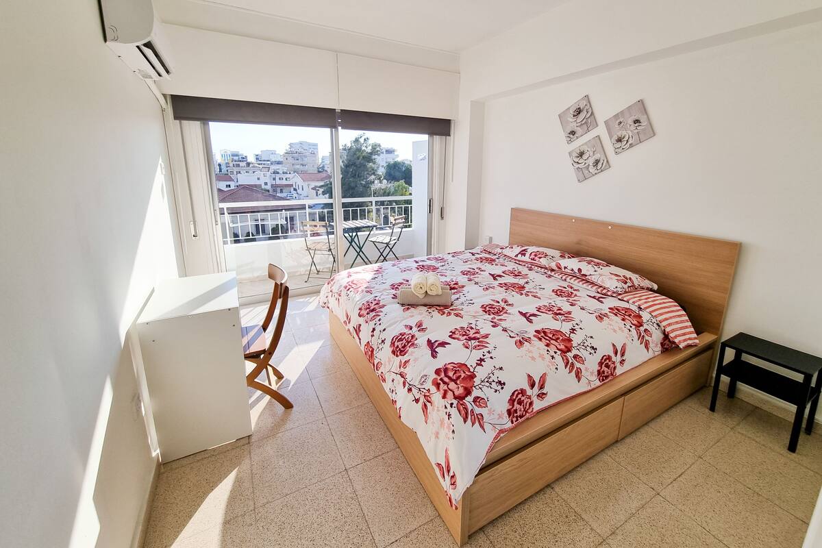 A bedroom with a bed, a desk, a chair, and a balcony with a view of buildings.