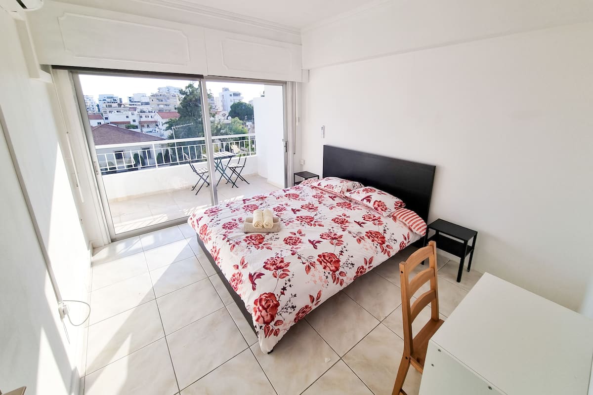 A bedroom with a bed, a chair, a table, and a balcony with a view of buildings.