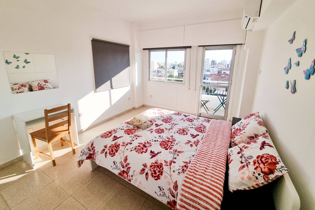 A bedroom with a bed, a desk, a chair, and a balcony with a view of the city.