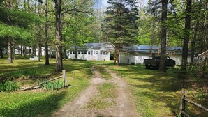 A rural property with a dirt path leading to a white building with a blue roof, surrounded by trees and a parked truck. - Summer fun in the Huron National Forest just minutes from the Au Sable River. (Glennie)