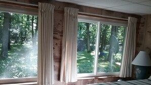 A bedroom with a large bed, two windows with curtains, a lamp, and a view of a forest. - Summer fun in the Huron National Forest just minutes from the Au Sable River. (Glennie)