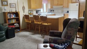 A living room with a sofa, a coffee table, a bookshelf, and a kitchen area with wooden cabinets and a refrigerator. - Summer fun in the Huron National Forest just minutes from the Au Sable River. (Glennie)