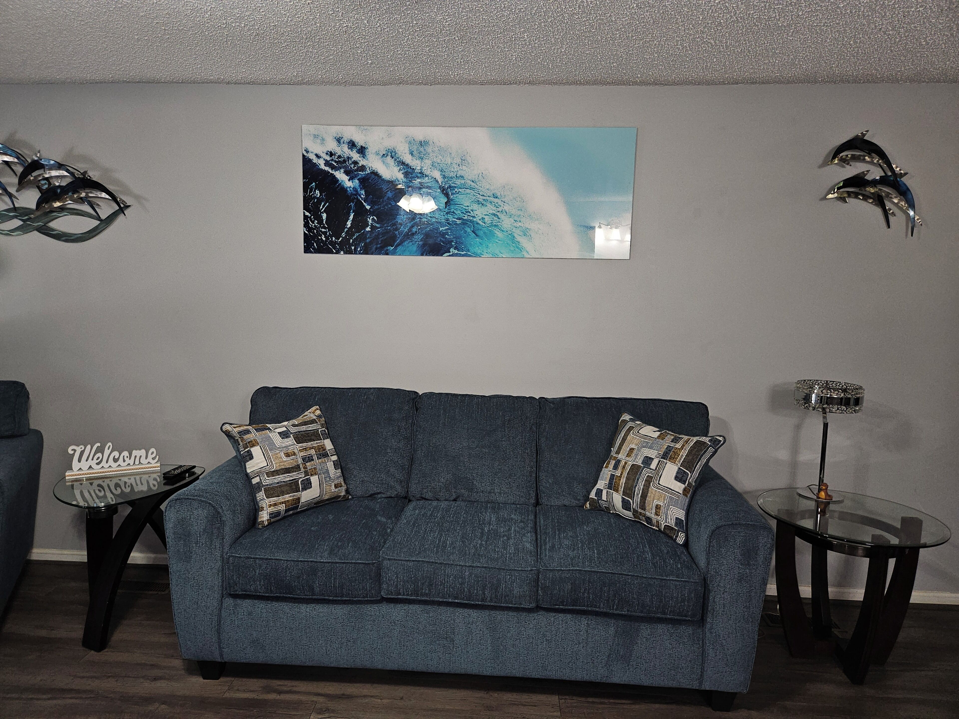 A living room with a dark blue sofa, two patterned throw pillows, a glass coffee table, a small side table, and a framed picture on the wall.