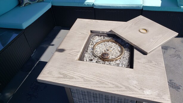 Un sofá modular con cojines estampados, una mesa cuadrada con aro metálico y tapa de madera, y un pequeño hogar con piedras de vidrio.