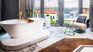 A modern bathroom with a freestanding bathtub, a small round table, and a sofa with a view of the city and river. - The Maestro Hotel (Istanbul)