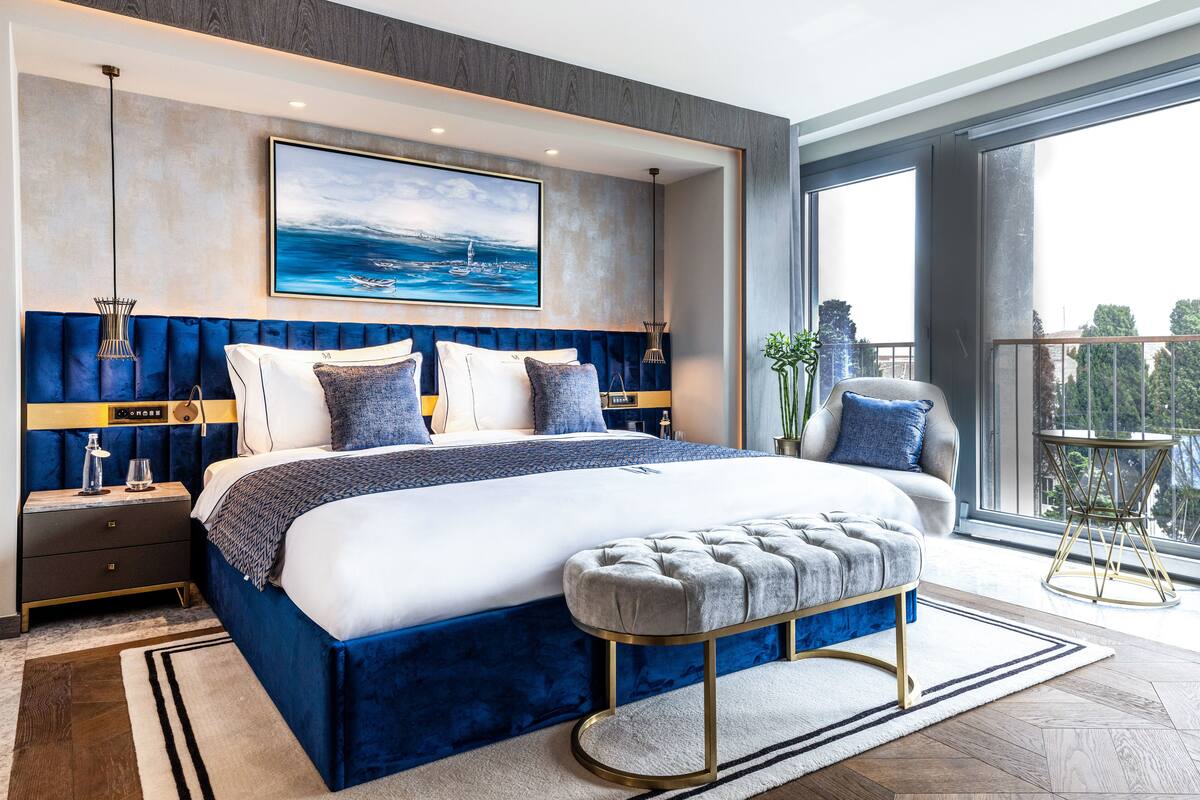 A modern bedroom with a large bed, a headboard with vertical lines, a bedside table, a bench, and a view of the outdoors through a large window.