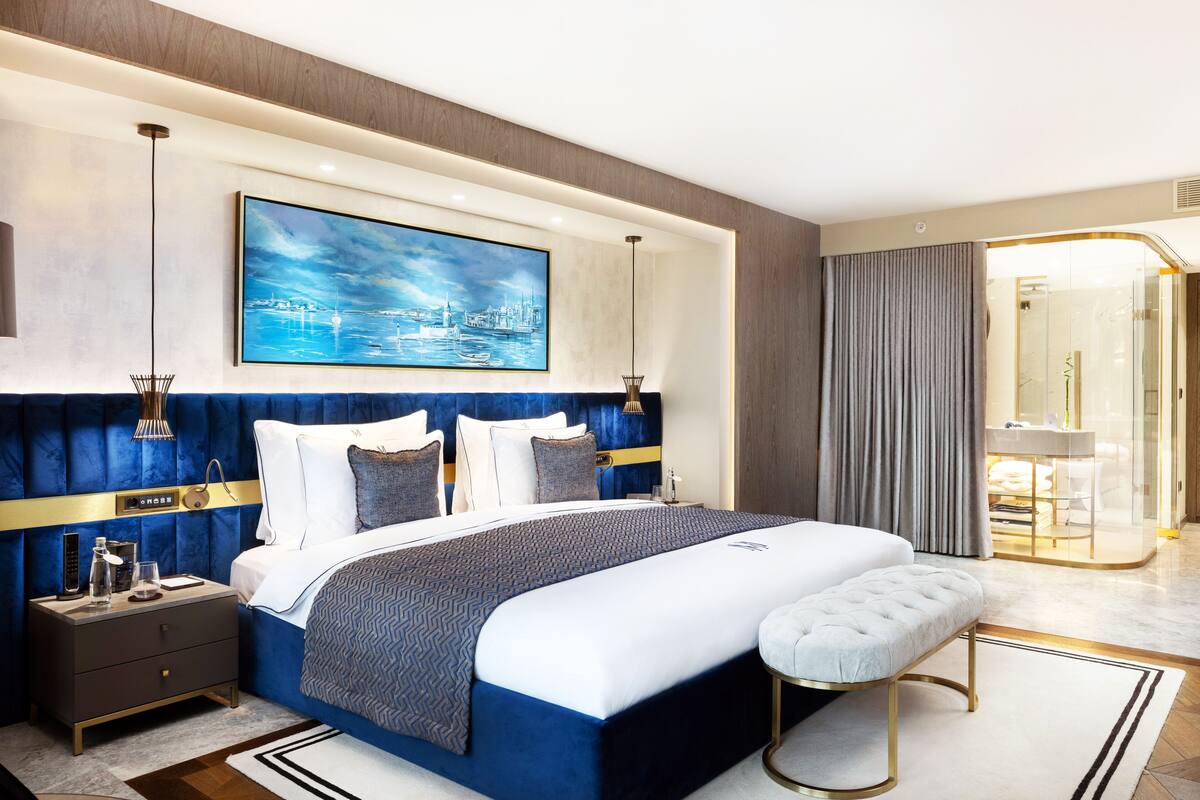 A modern hotel room with a large bed, a nightstand, a bench, a painting, and a glass-enclosed bathroom.
