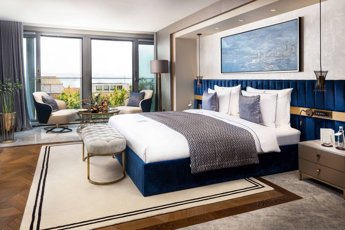 A modern hotel room with a large bed, a seating area with a table, and a view of the city through large windows.