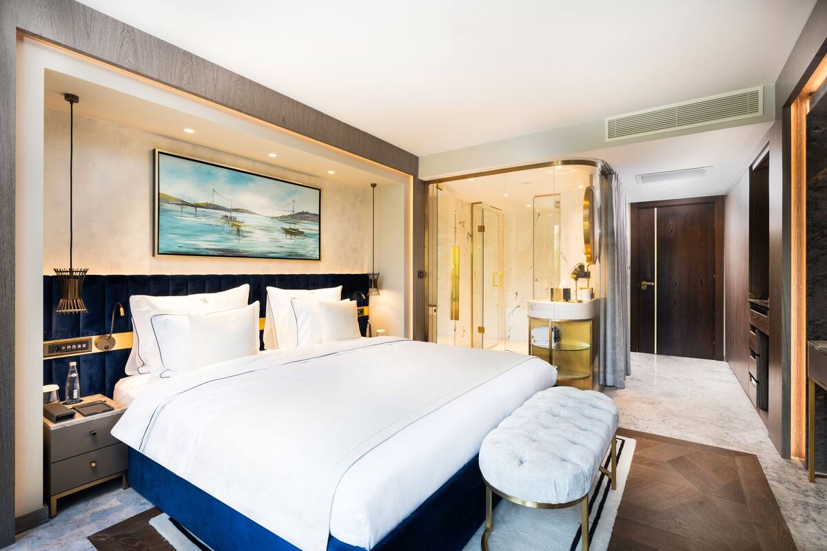 A modern hotel room with a large bed, a bedside table, a nightstand, a bathroom with a shower, and a large window.