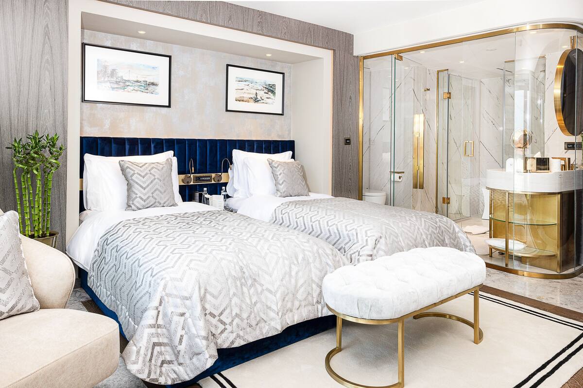 A modern hotel room with a large bed, a sitting area, a glass-enclosed shower, and a vanity with a mirror.