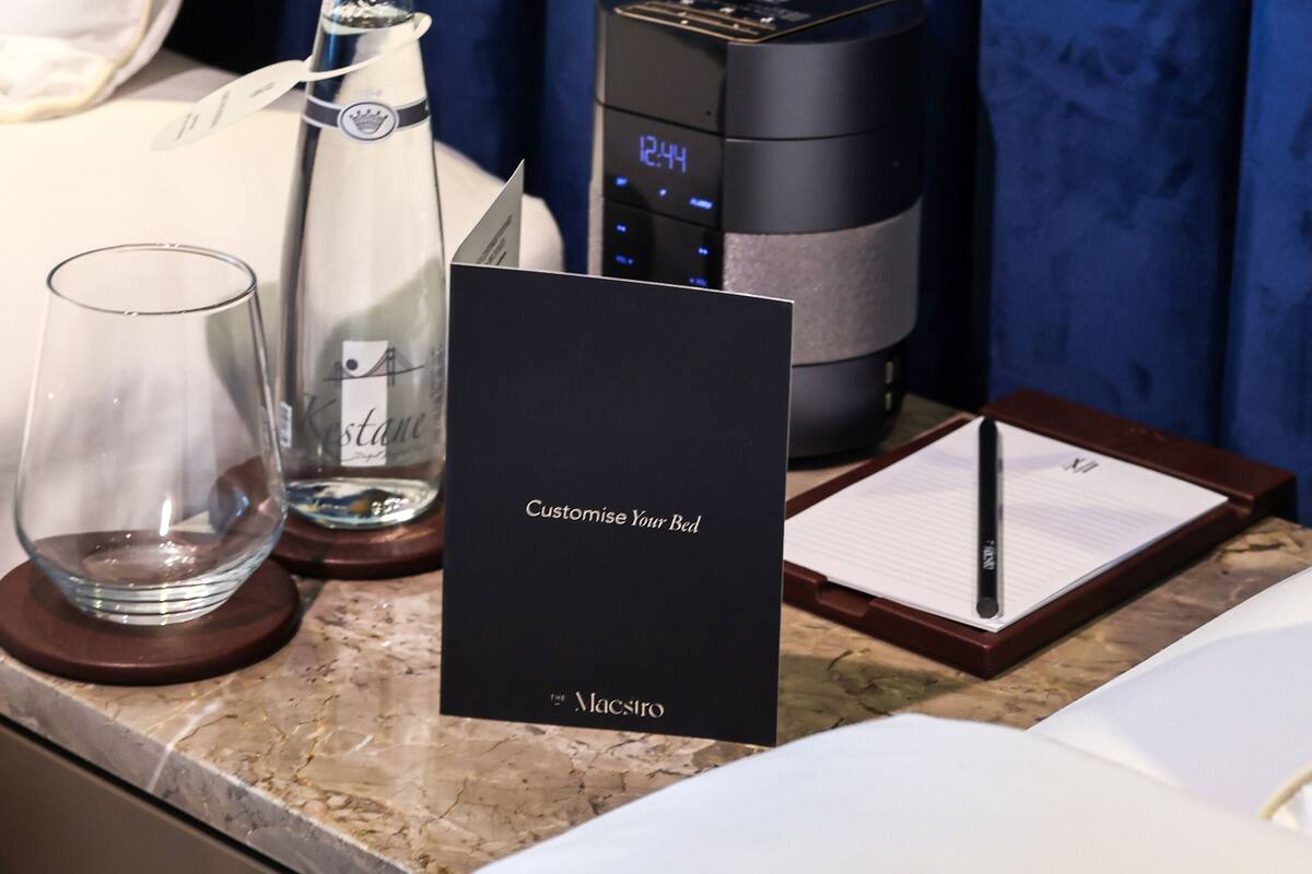 A hotel room with a bed, a bedside table, a bottle of water, a glass, a digital clock, and a bedside lamp.