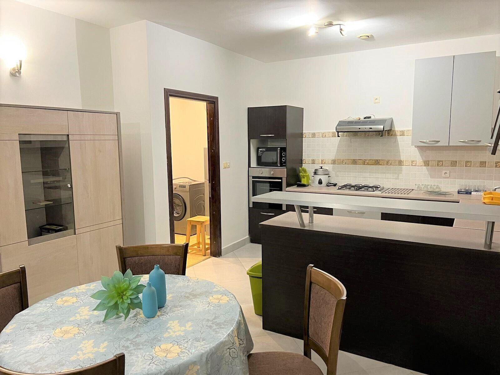A compact kitchen with a central island, a dining area with a table and chairs, and a laundry area visible through an open door.