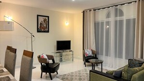 A modern living room with a flat-screen TV, a dining area with a table and chairs, and a large window with sheer curtains. - Appartements - Le Paradisier Ambatobe (Antananarivo)