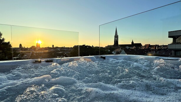Jacuzzi en la azotea con vista panorámica de la ciudad, incluyendo una aguja de iglesia, durante el atardecer.