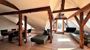 Un amplio dormitorio en el ático con techo de vigas de madera, una mesa de comedor con sillas, un sofá y un amplio ventanal con vistas al exterior.