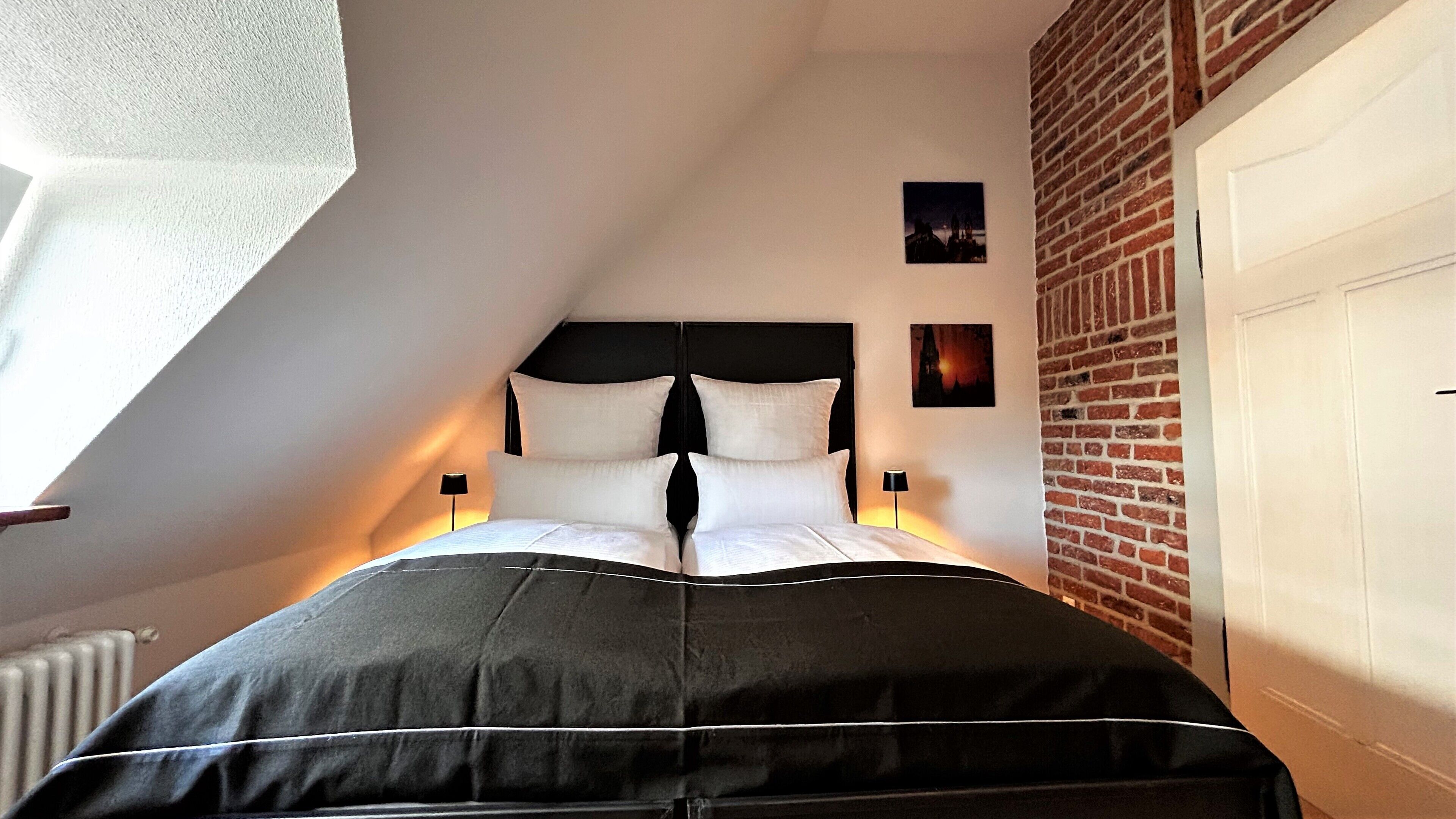 Un dormitorio con techo inclinado, cabecero negro, ropa de cama blanca y dos lámparas fijadas a la pared.