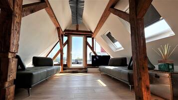 A loft bedroom with a sloped ceiling, wooden beams, a skylight, two sofas, a TV, and a small side table with a decorative item.