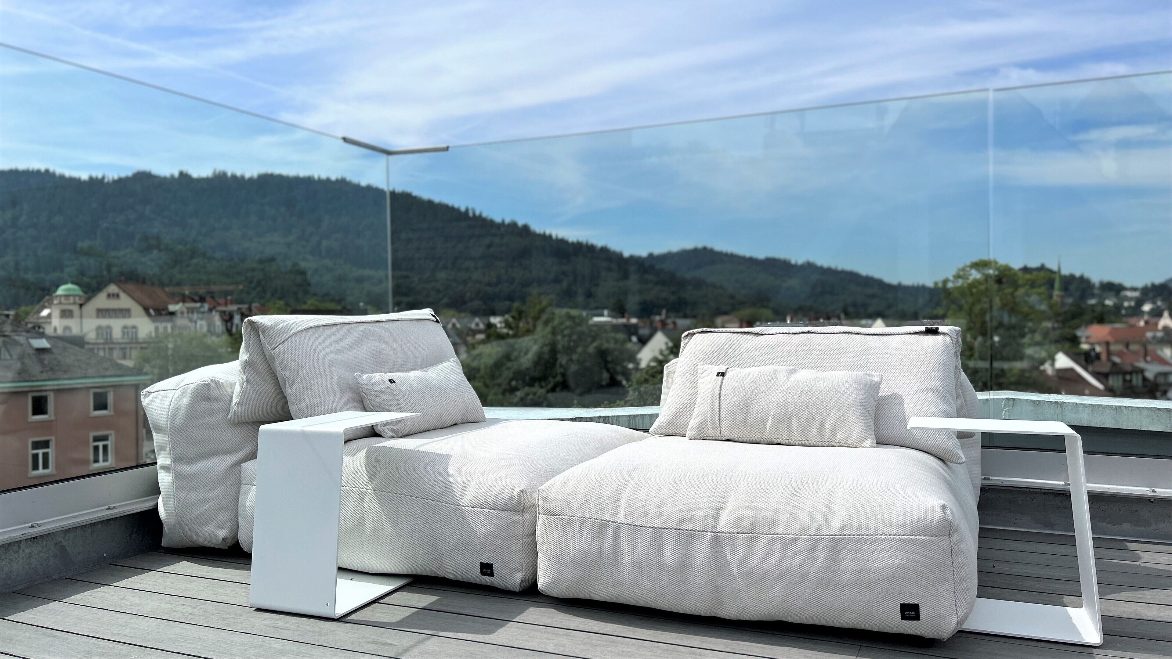 Un canapé blanc pour l’extérieur, placé sur un balcon, d’où l’on a une vue dégagée sur un paysage vallonné et des bâtiments en arrière-plan.