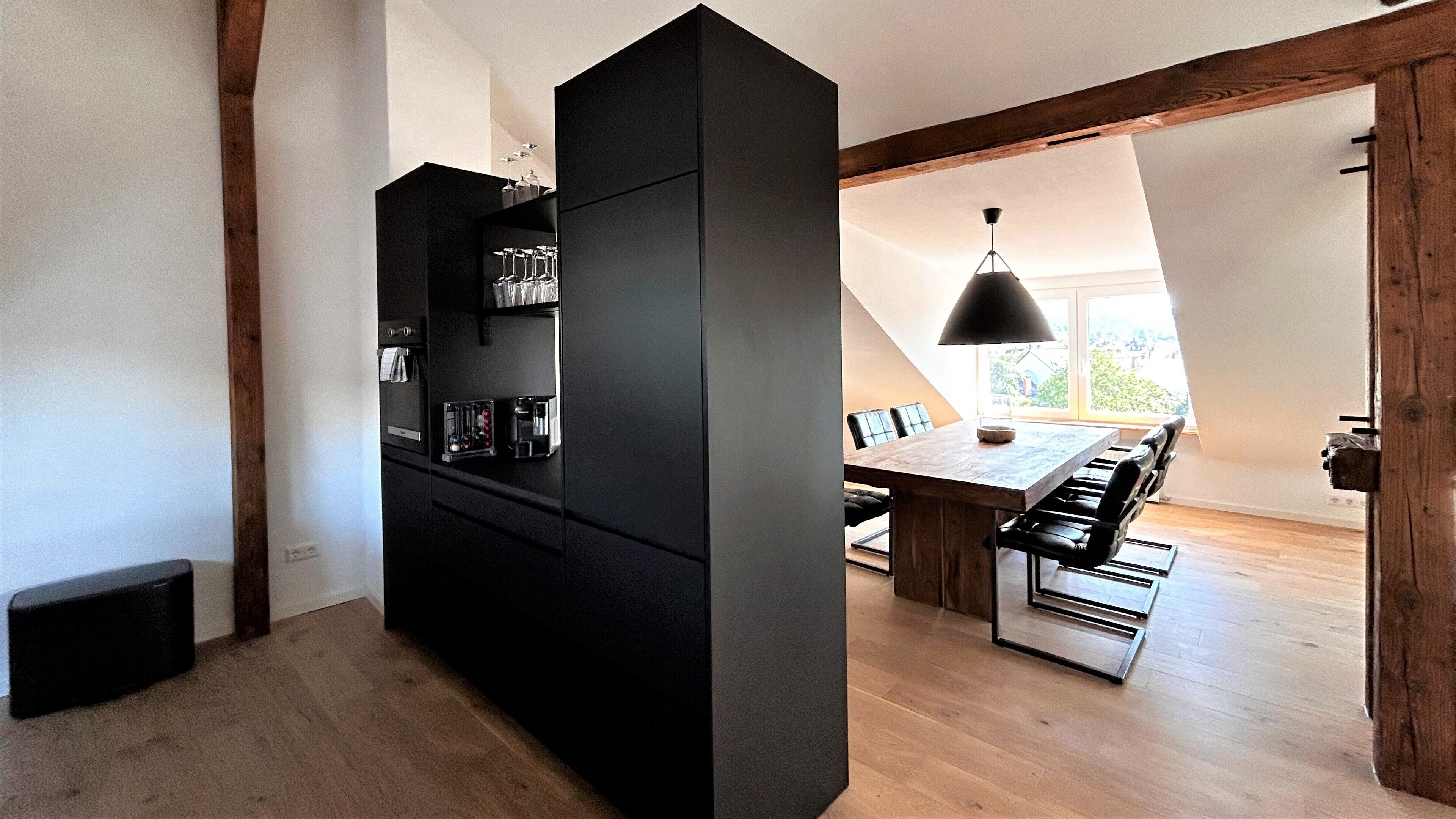 Une cuisine moderne avec des meubles de couleur noire, un coin repas avec une table et des chaises en bois, et une vue sur l’extérieur grâce à une grande fenêtre.