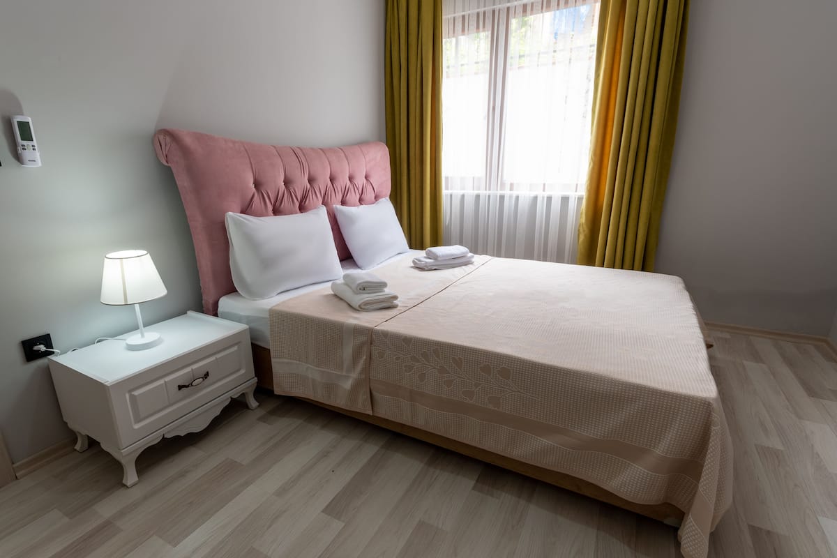Una cama bien hecha con cabecera rosa, almohadas blancas y colcha color beige. Hay una mesita de noche blanca con una lámpara y una toalla doblada. El cuarto tiene piso de madera clara y una ventana con cortinas amarillas.