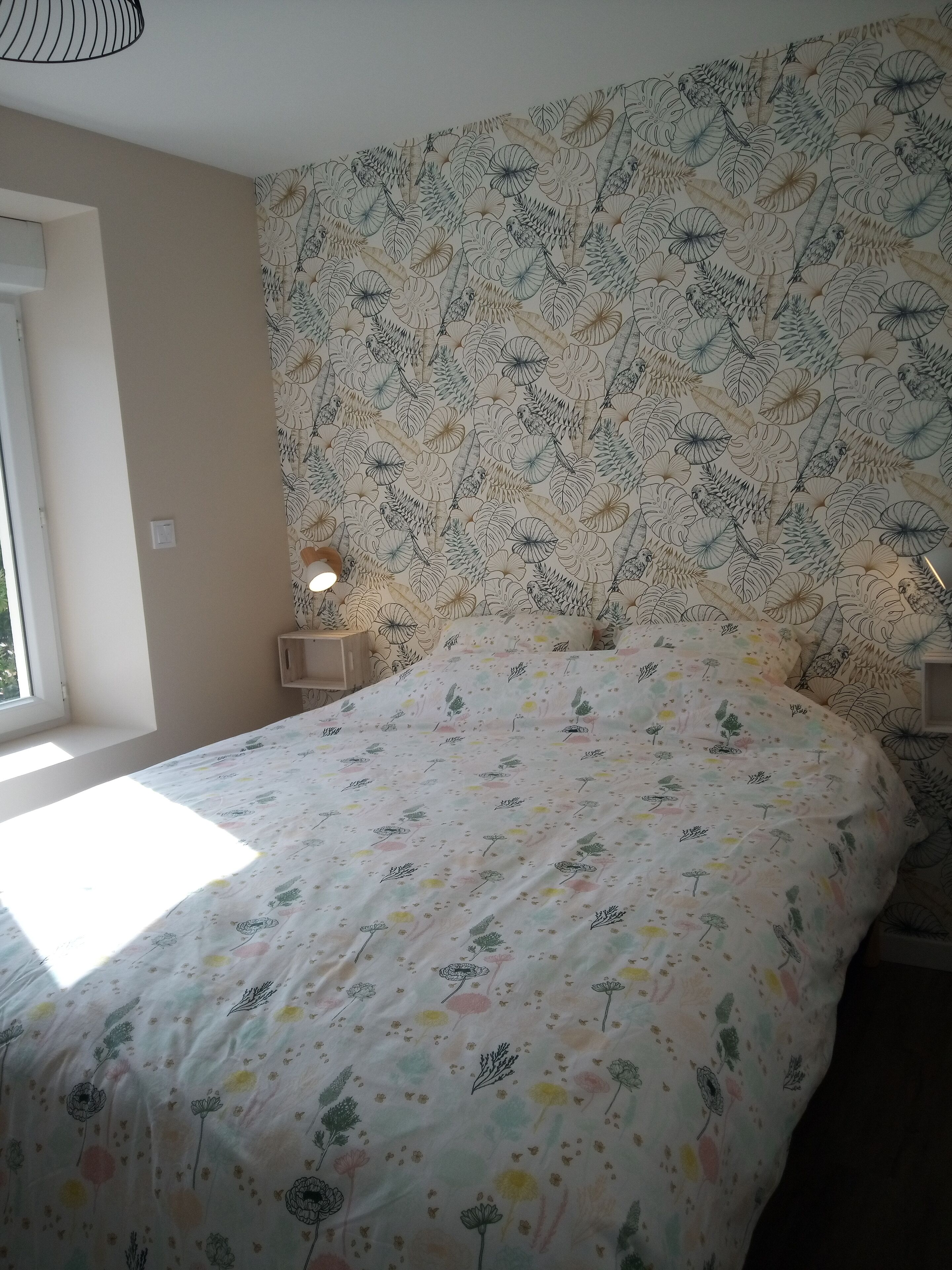 A bedroom with a floral-patterned bedspread, a window with natural light, and a wall with a leaf-pattern wallpaper.