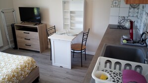 A compact hotel room with a bed, a desk with a chair, a television, a kitchenette, and a laundry basket.