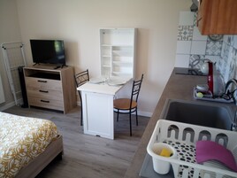 A compact hotel room with a bed, a desk with a chair, a television, a kitchenette, and a laundry basket.