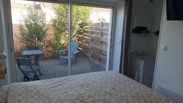 Quarto com cama, porta de vidro deslizante para um pátio com mesa e cadeiras, e banheiro pequeno com pia e espelho.