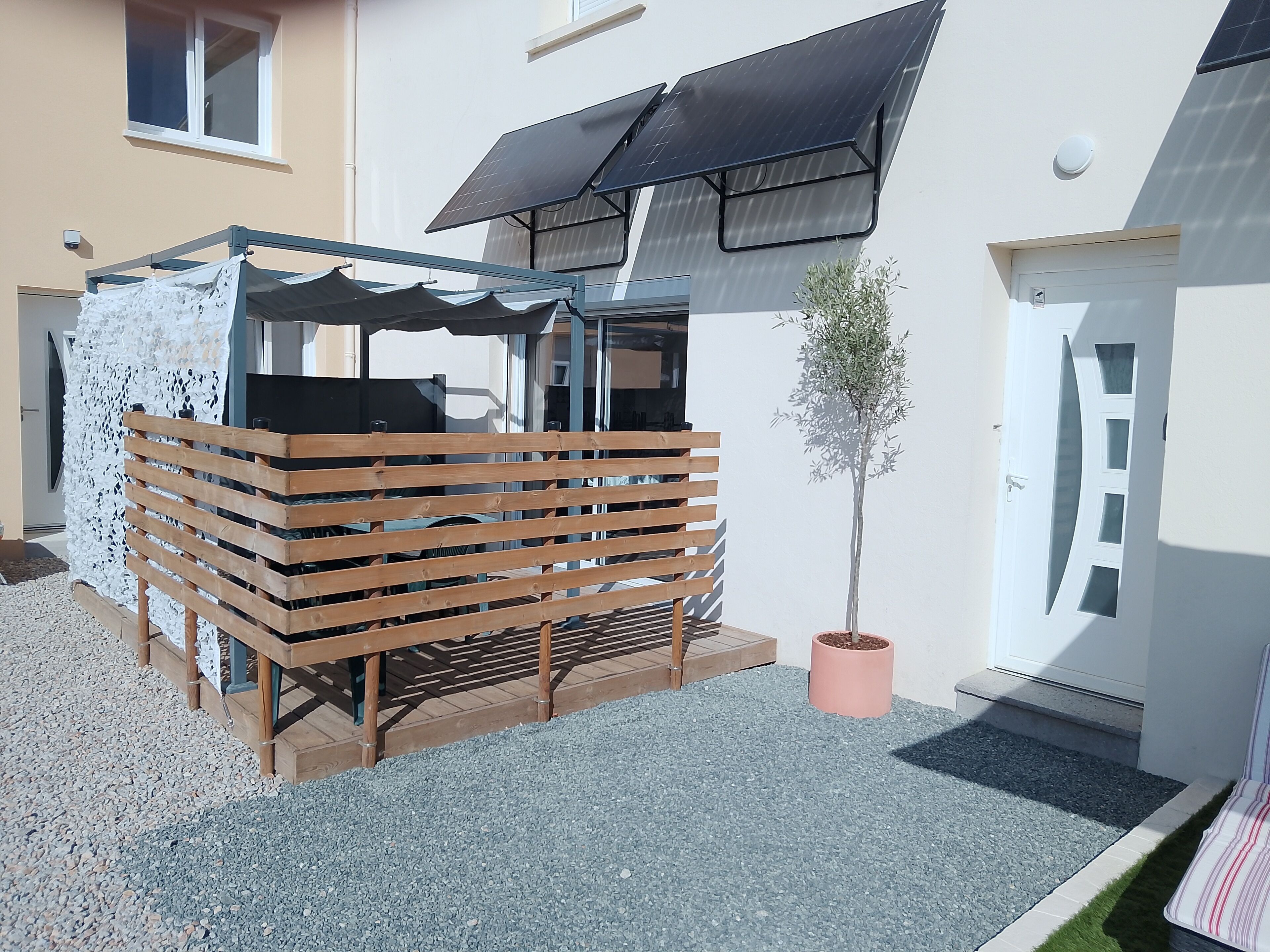 A covered outdoor seating area with a wooden fence, a potted plant, and a building with a solar panel.