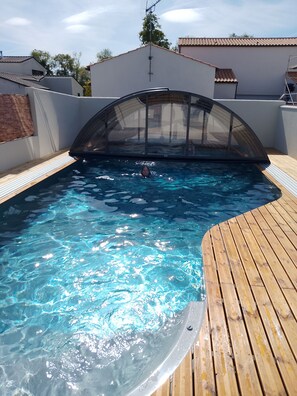 An outdoor swimming pool with a wooden deck, a covered area, and a clear blue sky.