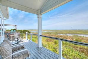 A covered outdoor deck with wicker furniture and a view of a marshy landscape. - Experience ocean views at 'West with the Night' in Oak Island, perfect for families. (Oak Island)