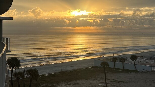 Vista sulla spiaggia con il sole che tramonta sull'oceano, palme e una passerella.