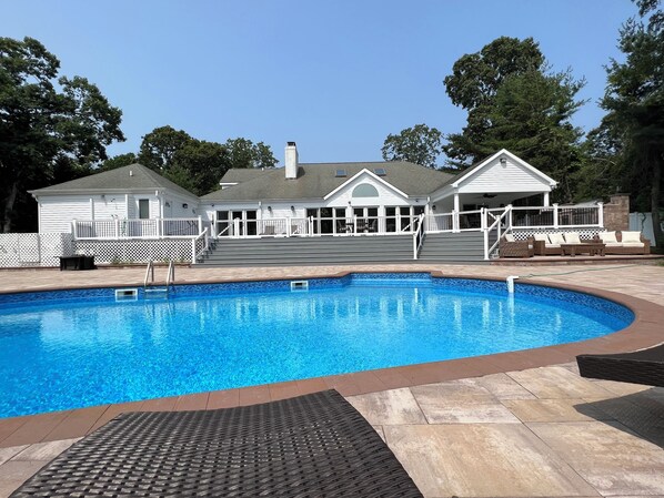 Ein Swimmingpool mit einer Steindeck, einer überdachten Terrasse und ein weißes Haus mit einem eingezäunten Deck im Hintergrund.