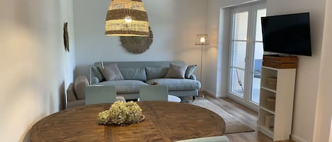 Een moderne woonkamer met een ronde houten eettafel, een lichtblauwe bank en een grote flatscreen televisie aan de muur.