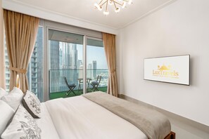 A hotel room with a large bed, a view of the city through floor-to-ceiling windows, a small balcony with a table and chairs, and a framed sign on the wall.