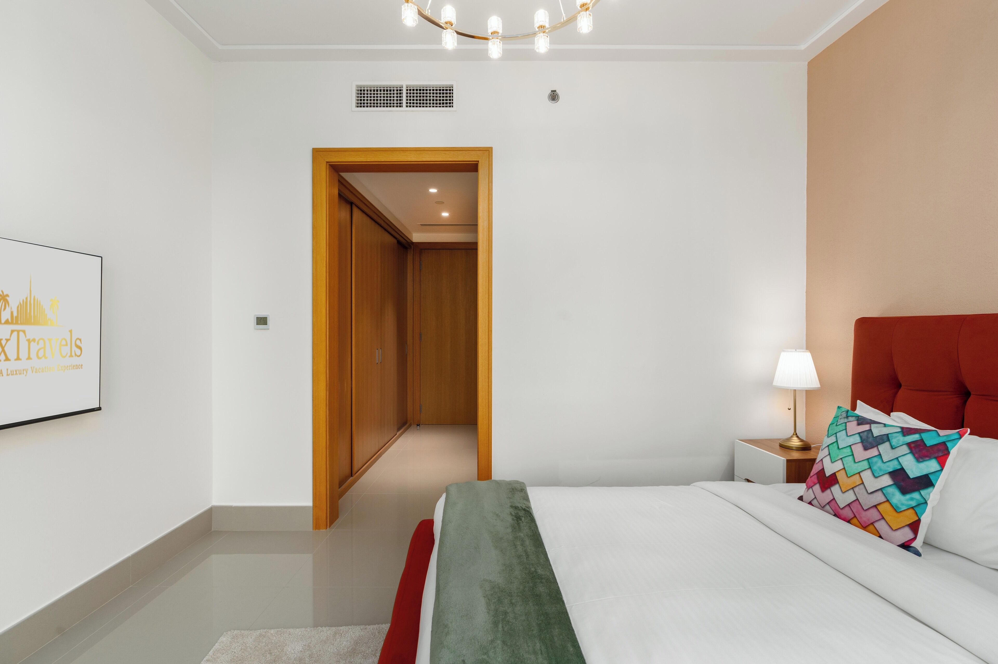 Una moderna camera d'albergo con un letto, un comodino, una lampada, un cuscino colorato, un quadro appeso alla parete e una porta in legno che dà su un corridoio.