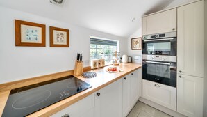 A modern kitchen with white cabinets, a black induction cooktop, and a built-in oven. There are framed pictures on the wall, a window with blinds, and a countertop with kitchen utensils. - Lower Farm Cottage -  Stalbridge Weston, Sherborne (Sturminster Newton, Dorset)