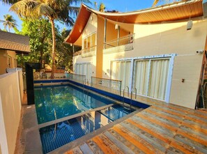 A swimming pool with a wooden deck, a covered balcony, and a white building with a garage.