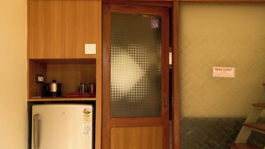 A compact room with a kitchenette, a bed with floral bedding, a wooden door with a frosted glass panel, and a small refrigerator. - Adrista Divine Cottages (Arambol)
