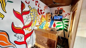 A room with a wooden reception desk, a television mounted on the wall, and colorful wall art. - Adrista Divine Cottages (Arambol)