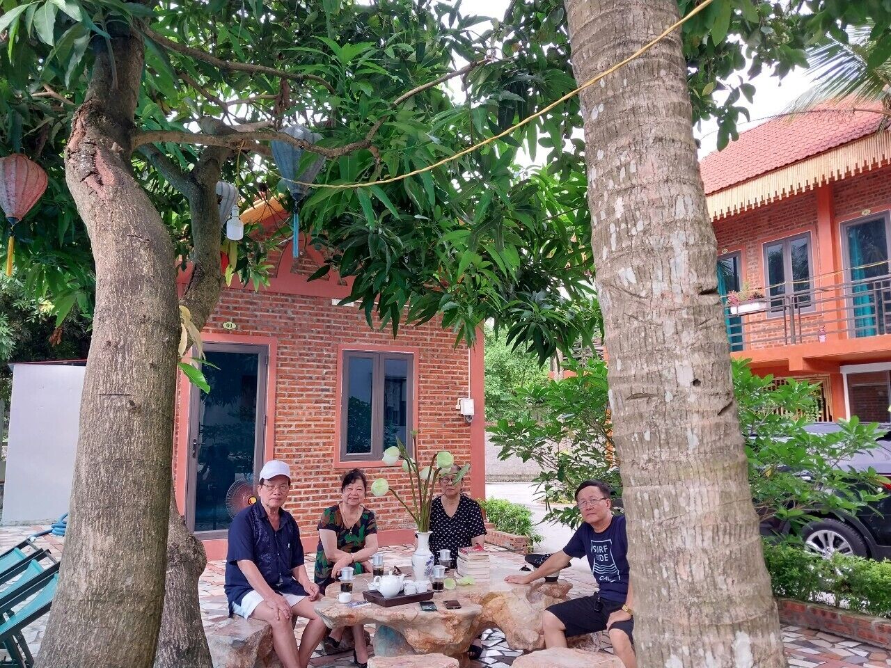 Eine Gruppe von Personen sitzt draußen an einem Tisch, im Hintergrund ist ein Backsteinhaus zu sehen.