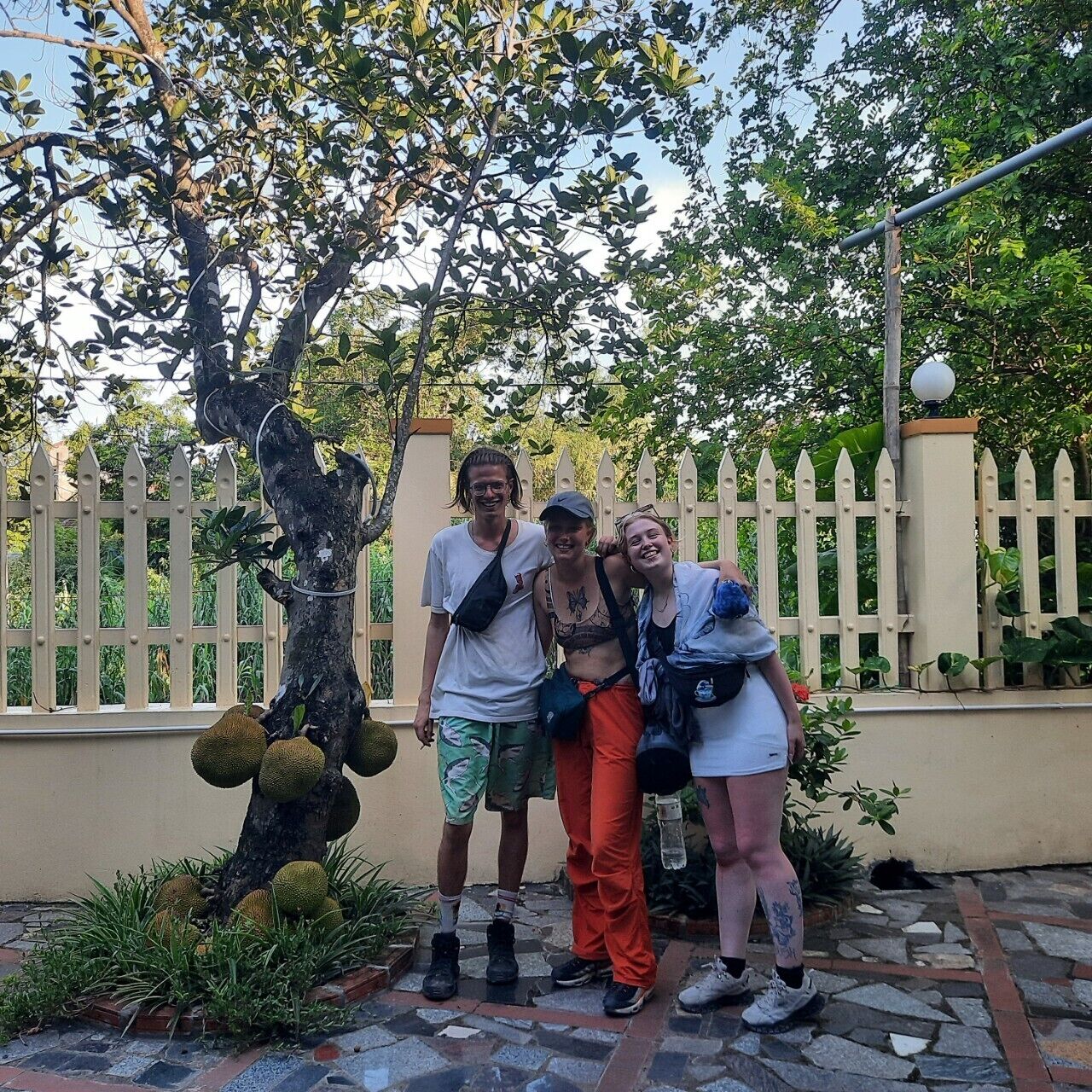 A group of people posing in front of a tree with large fruit, a white picket fence, and a lamp post in a garden setting.
