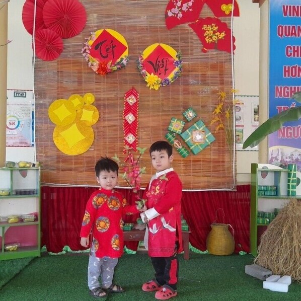 Dos niños con atuendo tradicional frente a un decorado adornado con elementos rojos y amarillos, posiblemente para un evento cultural.