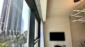 A modern living room with a large window, a flat-screen TV mounted on the wall, a small cabinet, and a unique ceiling light fixture. - LuxBnB Dubai Opera Act-1 Boulevard Views (Dubai)