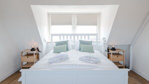 Een slaapkamer met een schuine wand, een groot raam, een bed met kussens, twee nachtkastjes met lampen, en houten nachtkastjes.