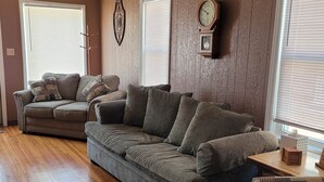Un salón con un sofá modular grande, un sofá más pequeño y un sillón estampado. Hay dos ventanas con persianas, un reloj de pared y un adorno colgante en la pared.