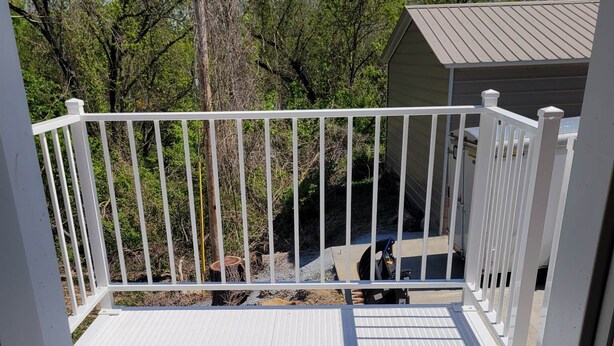 Una terraza con barandal blanco, escalera de metal y vista a árboles y un edificio al fondo.